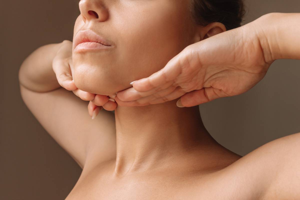 The image shows a young woman touching under the chin with hands massaging her face. The image represents the question of whether a midface lift can look stretched.